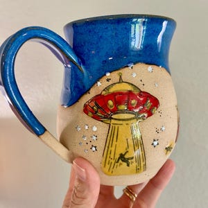 May include: A ceramic mug with a glossy blue upper section and a matte beige lower section. The beige area features a hand-painted yellow and red UFO with a beam of light shining down on a small figure, surrounded by small white star accents.
