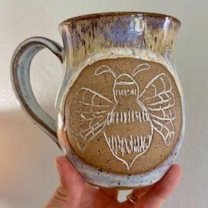 May include: A ceramic mug with a speckled tan body and a hand-carved bee design in white. The upper portion of the mug features a blue and brown drip glaze finish. It has a curved handle on the left side and a rounded shape with a wide rim.