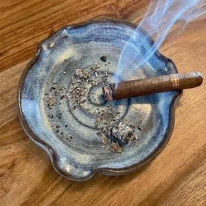 May include: A ceramic ashtray with a blue and white glaze holds a lit cigar, emitting wisps of smoke. The ashtray has a unique, organic shape and is filled with ash. The cigar is labeled "COHIBA".