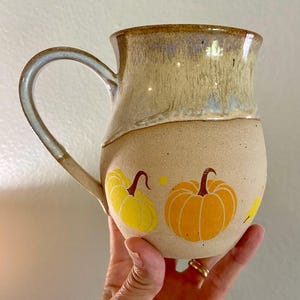 May include: A ceramic mug with a speckled beige base and a smooth, glossy tan glaze on the upper portion. The lower section features a hand-painted design of two orange and yellow pumpkins with brown stems, accented by small yellow dots and a leaf.