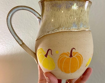 May include: A ceramic mug with a speckled beige base and a smooth, glossy tan glaze on the upper portion. The lower section features a hand-painted design of two orange and yellow pumpkins with brown stems, accented by small yellow dots and a leaf.