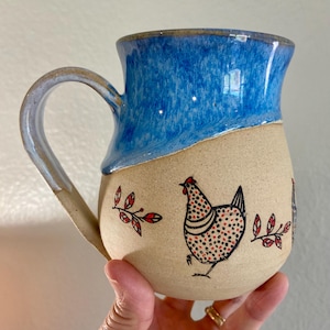 May include: A handmade ceramic mug with a blue glazed top and a tan base. The base features a hand-drawn chicken design with red and black accents, along with red leaf accents. The mug has a curved handle.