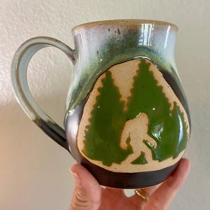 May include: A ceramic mug with a curved handle featuring a glazed finish in shades of blue, green, and dark brown. The front displays a carved, unglazed tan section depicting a silhouette of a creature resembling Bigfoot among pine trees.