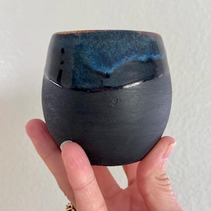 May include: A black ceramic cup with a dark blue glaze on the top half. The cup is round and has a wide opening.