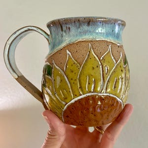 May include: A ceramic mug with a speckled tan base featuring a hand-carved sunflower design with yellow petals and a brown center. The upper portion of the mug and the handle are glazed in a glossy light blue, with a smooth, curved rim and handle.