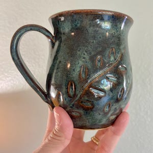 May include: Handmade ceramic mug with a teal and brown glaze. The mug features an embossed leaf design and a curved handle. The interior and rim have a darker brown color. A unique piece for enjoying coffee or tea.