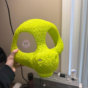 May include: A neon green, textured mask with large eye openings and a snout. The mask is held up in front of a sewing machine and a window. The mask appears to be 3D printed.