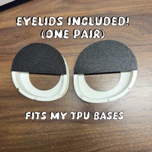 Pode incluir: Um par de componentes circulares de pl&aacute;stico branco com coberturas semicirculares pretas texturizadas na metade superior. O texto EYELIDS INCLUDED! (ONE PAIR) e FITS MY TPU BASES &eacute; vis&iacute;vel perto de um dispositivo Bambu Lab.