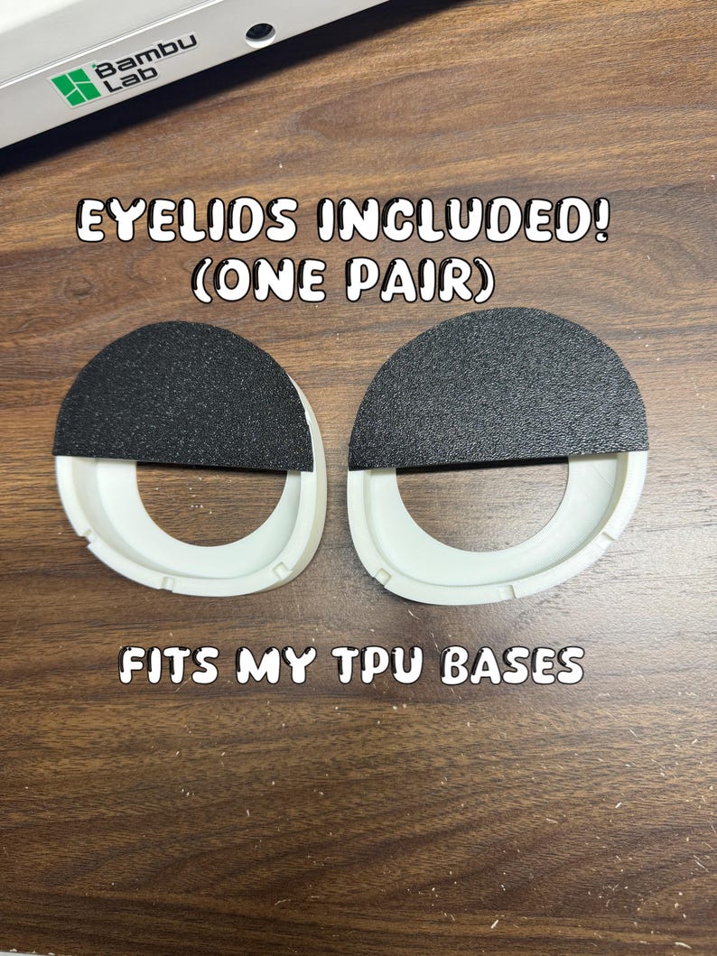Pode incluir: Um par de componentes circulares de pl&aacute;stico branco com coberturas semicirculares pretas texturizadas na metade superior. O texto EYELIDS INCLUDED! (ONE PAIR) e FITS MY TPU BASES &eacute; vis&iacute;vel perto de um dispositivo Bambu Lab.