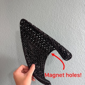 Könnte beinhalten: Ein schwarzes, 3D-gedrucktes, dreieckiges Ohr-Accessoire mit einer texturierten, gitterartigen Oberfläche. Das Objekt hat eine geschwungene Basis und eine spitze Form, mit einem roten Pfeil, der auf eine Öffnung mit dem Text Magnet holes! zeigt.