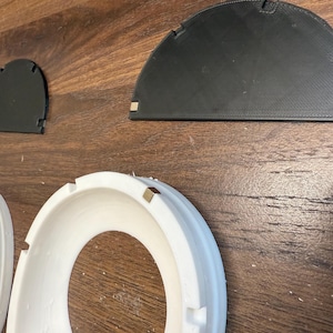 Pode incluir: Um anel circular branco com um pequeno cubo met&aacute;lico embutido na borda, acompanhado por duas pe&ccedil;as planas semicirculares pretas, cada uma com um pequeno cubo met&aacute;lico em um canto, projetadas para montagem ou uso modular.
