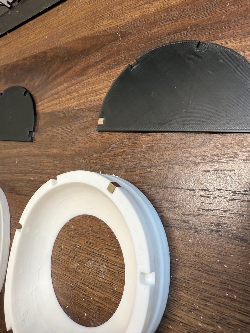 Pode incluir: Um anel circular branco com um pequeno cubo met&aacute;lico embutido na borda, acompanhado por duas pe&ccedil;as planas semicirculares pretas, cada uma com um pequeno cubo met&aacute;lico em um canto, projetadas para montagem ou uso modular.