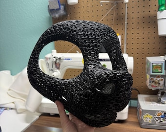 Pode incluir: Uma base de máscara de animal preta impressa em 3D com uma estrutura texturizada semelhante a uma rede, com grandes aberturas para os olhos e um focinho proeminente. A máscara tem um interior oco e uma forma arredondada com um padrão de filamento detalhado.