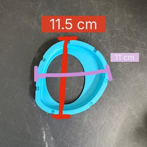 K&ouml;nnte beinhalten: Ein hellblauer, kreisf&ouml;rmiger, 3D-gedruckter Kunststoffrahmen mit einer zentralen &Ouml;ffnung. Das Objekt weist ein vertikales Ma&szlig; von 11,5 cm und ein horizontales Ma&szlig; von 11 cm auf, wie durch die roten und violetten Linien im Bild dargestellt.