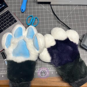 May include: A pair of plush animal-style paw gloves. One glove features white faux fur with light blue paw pads, while the other is covered in dark purple faux fur with white paw pads. Both gloves have dark grey cuffs at the wrist openings.