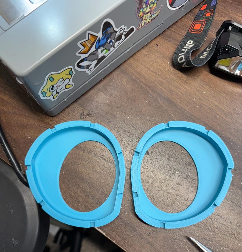 K&ouml;nnte beinhalten: Zwei identische, leuchtend blaue, kreisf&ouml;rmige 3D-gedruckte Kunststoffrahmen mit asymmetrischem Innenausschnitt und kleinen Kerben am Au&szlig;enrand. Diese Bauteile haben eine glatte, matte Oberfl&auml;che und sind mit einem erh&ouml;hten Rand versehen.