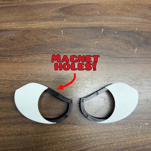 Könnte beinhalten: Zwei 3D-gedruckte augenförmige Komponenten mit einer weißen Außenhülle und einem schwarzen Innenrahmen. Jedes Teil enthält kleine quadratische Vertiefungen, die mit dem roten Text MAGNET HOLES! und einem roten Pfeil markiert sind.
