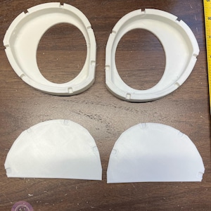 K&ouml;nnte beinhalten: Zwei wei&szlig;e, 3D-gedruckte ovale Rahmen mit gekerbten Kanten und zwei passende wei&szlig;e halbkreisf&ouml;rmige Eins&auml;tze. Ein gelbes Ma&szlig;band ist auf der rechten Seite sichtbar und zeigt, dass die Objekte etwa 7 bis 10 cm breit sind.