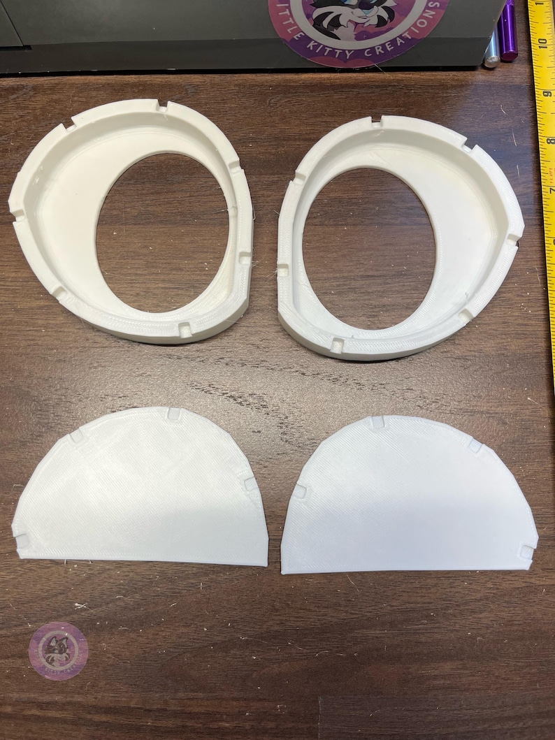 K&ouml;nnte beinhalten: Zwei wei&szlig;e, 3D-gedruckte ovale Rahmen mit gekerbten Kanten und zwei passende wei&szlig;e halbkreisf&ouml;rmige Eins&auml;tze. Ein gelbes Ma&szlig;band ist auf der rechten Seite sichtbar und zeigt, dass die Objekte etwa 7 bis 10 cm breit sind.