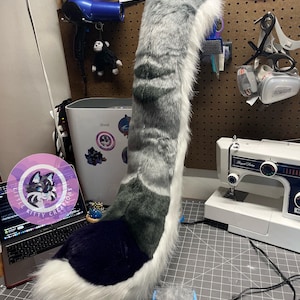 May include: A long, plush faux fur tail featuring a gray and white striped pattern with a dark purple tip. The tail has a thick, soft texture and is positioned upright on a gray cutting mat next to a sewing machine and a blue grooming brush.