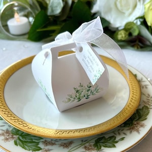 May include: A white wedding favour box with a handle, adorned with green floral designs, rests on a gold-edged plate. A white ribbon and tag with text reading "Thank you for sharing Neil and Jane's Wedding Day" are attached.