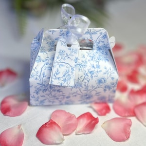 May include: A small, square gift box with a blue floral pattern and a white tag tied with a sheer ribbon. Pink rose petals are scattered around the box on a white surface. The box has a handle.