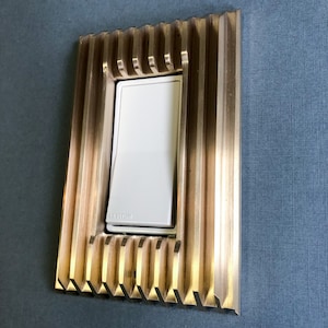 May include: A white light switch with the word "LEVITON" printed on it, surrounded by a gold metal frame with a decorative, ribbed design.