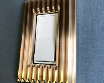 May include: A white light switch with the word "LEVITON" printed on it, surrounded by a gold metal frame with a decorative, ribbed design.