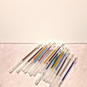 May include: A collection of clear plastic pens with various ink colors, including blue, brown, yellow, and green. The pens are arranged on a light-colored surface, with a white background.