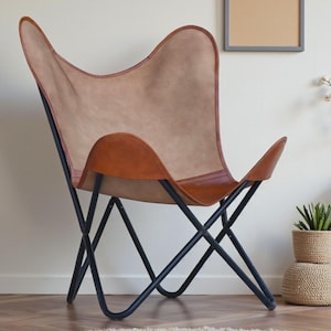 May include: A butterfly chair with a tan leather seat and brown leather trim. The chair has a black metal frame and is set against a neutral wall. Decorative items like a framed picture and a woven vase are in the background.