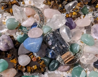 Può includere: Una varietà di cristalli e pietre, tra cui quarzo chiaro, quarzo rosa, occhio di tigre, ametista e agata blu. I cristalli sono disposti in una ciotola o un cestino, e c'è una piccola bottiglia di vetro piena di sabbia nera.