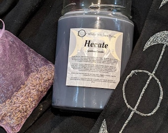 May include: A dark gray glass candle jar with a white label that reads "White Witches Brew Hecate Goddess Candle". The label includes a pentagram symbol and text about the Greek goddess Hecate. The candle is sitting on a black fabric with a silver embroidered design.