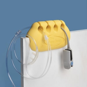 May include: A yellow plastic device labelled "The Venta Clasp" with slots, holding clear tubing and a grey sensor. The device is mounted on a white surface against a blue background. The tubing has a nasal cannula.