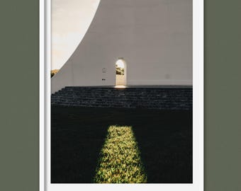 Pode incluir: Fotografia emoldurada com uma cena arquitetônica minimalista. Uma parede branca curva com uma porta é colocada contra um céu brilhante. Uma área retangular de grama ensolarada contrasta com o primeiro plano escuro.