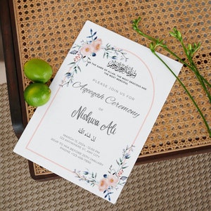 Puede incluir: Una tarjeta de invitaci&oacute;n blanca con dise&ntilde;os florales rosas y azules. La tarjeta anuncia una ceremonia Ageezah para Nishwa Ali el 13 de marzo de 2024 a las 12:00 PM. La fecha l&iacute;mite de respuesta es el 25 de enero. La tarjeta incluye la frase &aacute;rabe "إنْ شَاءَ اللهُ" que se traduce como "Si Dios quiere."