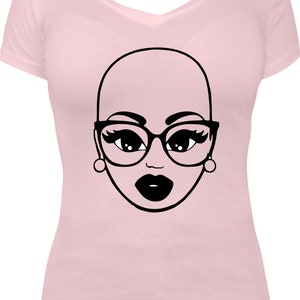 May include: A light pink V-neck t-shirt featuring a black and white graphic of a bald face wearing glasses, earrings, and dark lipstick. The graphic is centered on the front of the shirt.