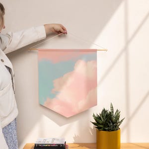 May include: A fabric wall hanging banner featuring a printed design of soft, fluffy pink clouds against a light blue sky. The banner is suspended from a horizontal wooden dowel with a thin white string attached to the ends for hanging.