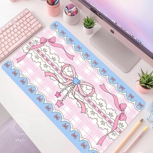 May include: A rectangular desk mat featuring a light pink and white plaid pattern, decorated with a scalloped blue border, white lace trim, and pink ribbons. A central pink bow is accented with a blue heart charm and dangling star charms.