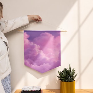 May include: A fabric wall banner featuring a printed image of fluffy clouds in shades of purple and pink. The banner has a pointed bottom edge and is suspended from a horizontal wooden dowel by a thin white string attached to both ends.