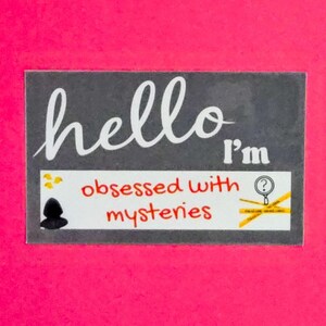 May include: A rectangular sign with the words "hello I'm obsessed with mysteries" in white and red lettering. The sign includes a magnifying glass and crime scene tape on a white background, set against a pink backdrop.