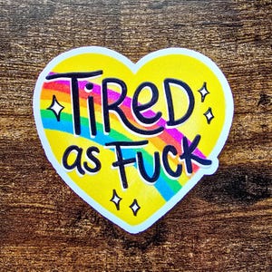 May include: A heart-shaped sticker with a yellow background and the words "Tired as Fuck" in black, outlined in white. The word "Tired" is above a rainbow stripe, and the sticker has four small star-shaped accents.