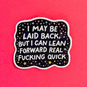 May include: A black sticker with a white border and the text "I MAY BE LAID BACK, BUT I CAN LEAN FORWARD REAL FUCKING QUICK" in white. The sticker has a starry design and is set against a bright pink background.