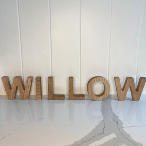 May include: Wooden letters spelling out the name "WILLOW" in a natural wood color. The letters are arranged in a line on a white countertop.