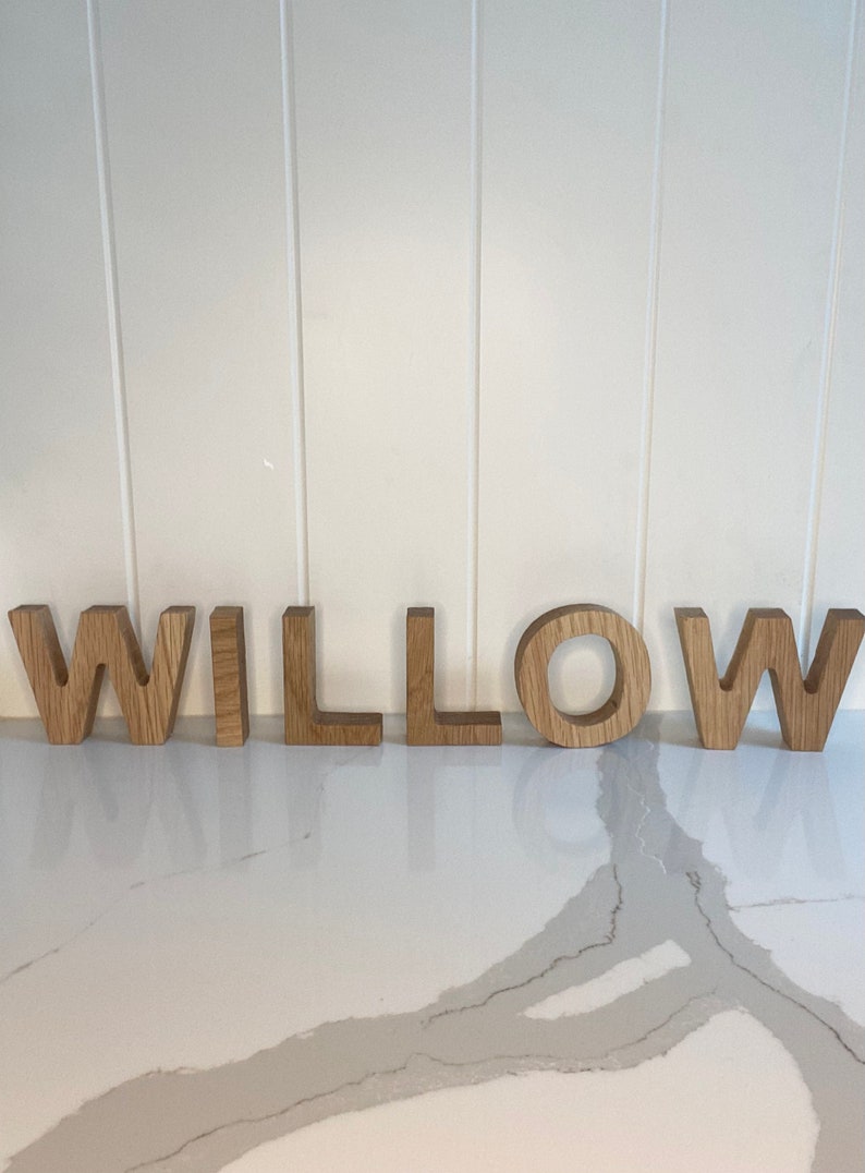 May include: Wooden letters spelling out the name "WILLOW" in a natural wood color. The letters are arranged in a line on a white countertop.