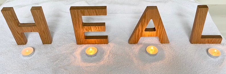 May include: Wooden letters spelling the word "HEAL" are arranged on a white surface. Four small, lit candles are placed in front of each letter.