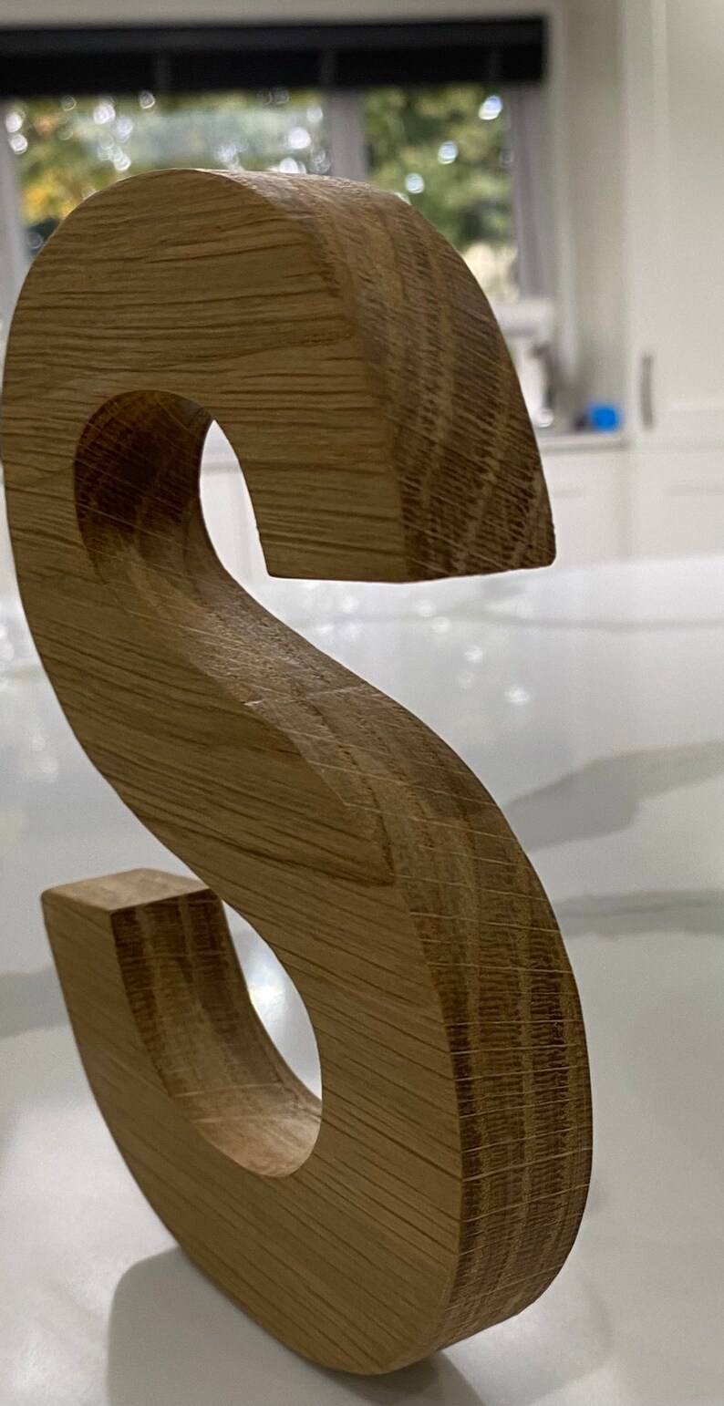 May include: A wooden letter S, crafted from light brown oak wood. The letter is freestanding and has a smooth, polished finish.