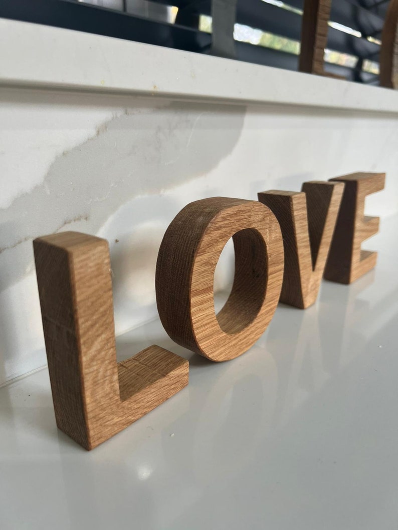 May include: Wooden letters spelling out the word "LOVE" in a light brown color. The letters are standing on a white surface.