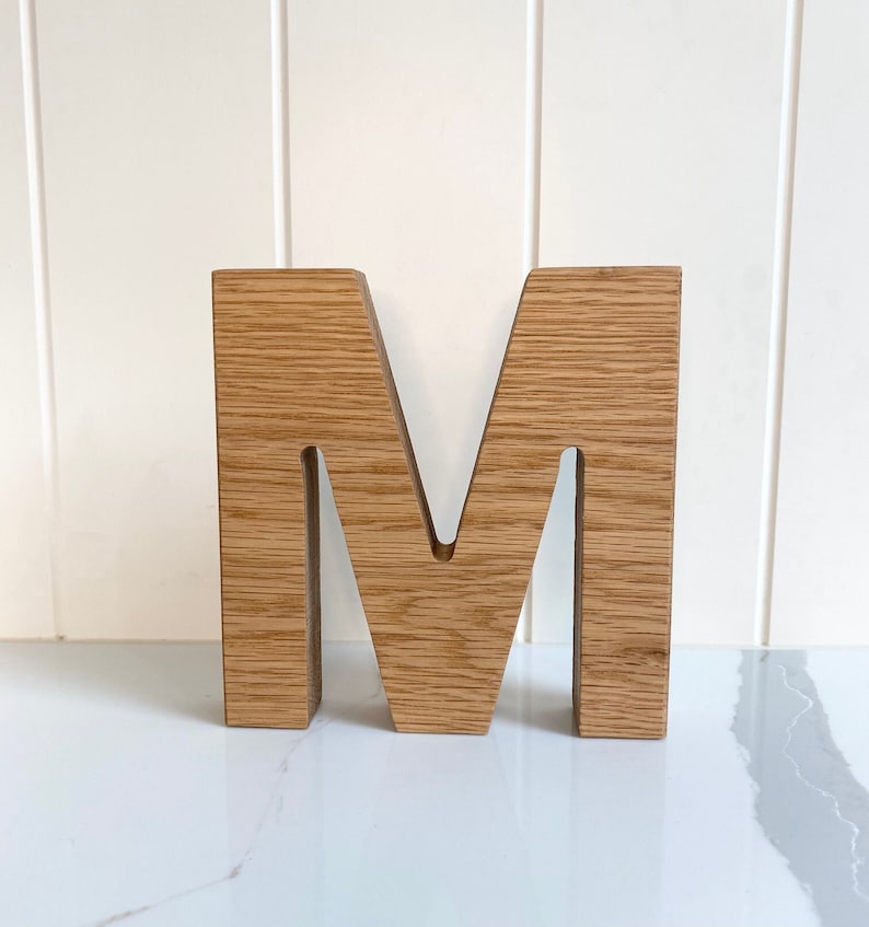 May include: A wooden letter M, standing upright on a white surface. The letter is made of light brown wood with a natural grain pattern.
