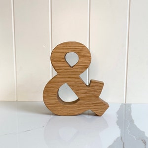 May include: Wooden ampersand symbol, carved from light brown wood. The ampersand is standing on a white surface.