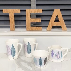 May include: A set of four ceramic mugs and a creamer with a leaf design in shades of purple, blue, and white. The mugs and creamer are on a white countertop with the word "TEA" spelled out in wooden letters.